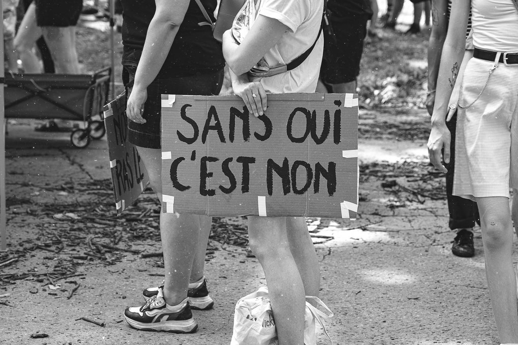 A group of people gather outdoors, with one person holding a sign that reads “Sans oui c'est non.” The scene is in black and white.