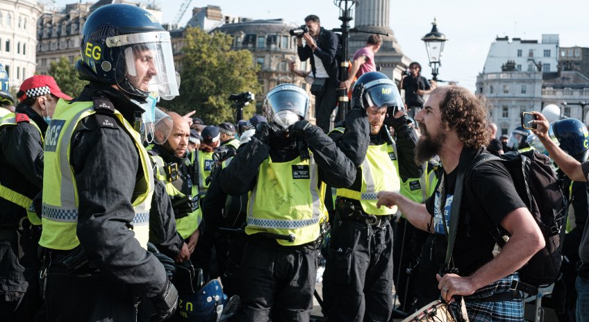 Police officers in protective gear and yellow vests face a group of protesters. Some protesters are filming the scene while others are arguing. Municipal buildings and a monument are visible in the background.