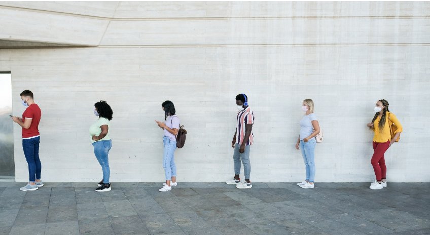 Sechs Personen stehen mit Abstand zueinander an einer hellen Wand. Sie sind in Freizeitkleidung und tragen Masken; eine Person hat einen Kopfhörer aufgesetzt, zwei Personen blicken auf ihre Smartphones.