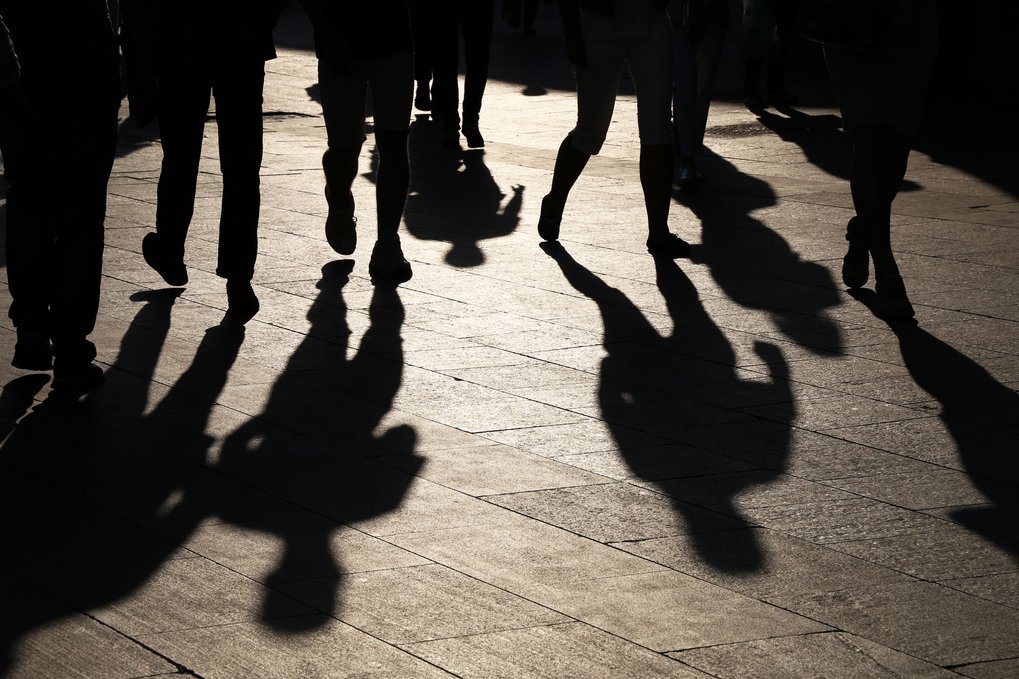 Silhouettes of pedestrians casting shadows on a sunlit sidewalk as they walk at dusk.
