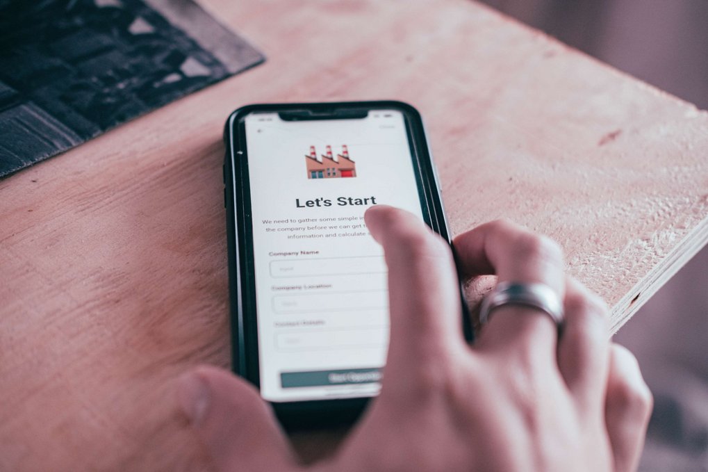 Close-up of a smartphone; the screen displays the text “Let's Start” with a form requesting company-related data. A person clicks on it with her/his finger.
