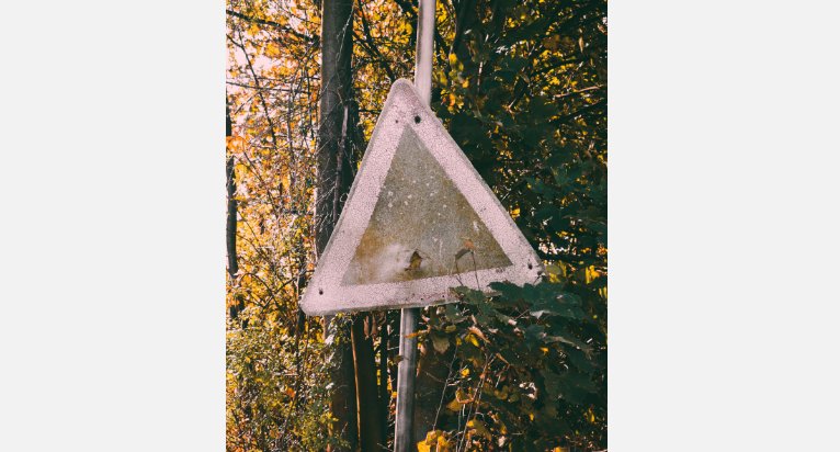 Altes, verrostetes, Verkehrsschild in Dreiecksform, das in einem herbstlichen Waldstück steht.