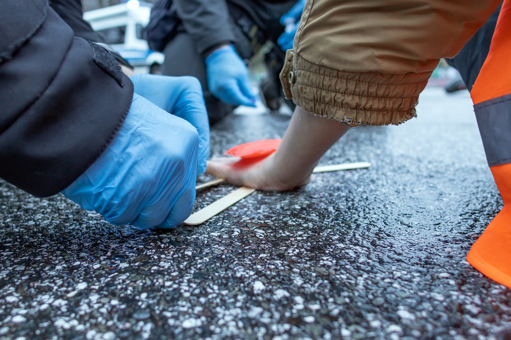 Klimakleber: Eine Person, die eine orangene Warnweste trägt, hat ihre Hand am Asphalt festgeklebt und andere Personen (Feuerwehr?) mit blauen Schutzhandschuhen versuchen, die Hand vom Asphalt zu lösen. 