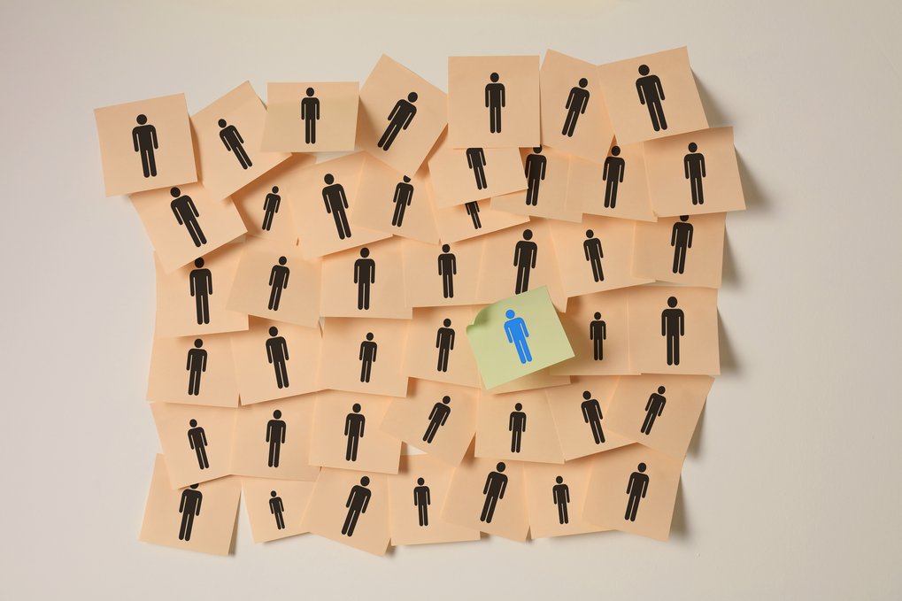 Numerous beige notes (sticky notes) with black figures are attached to a wall, with one green note with a blue figure standing out.