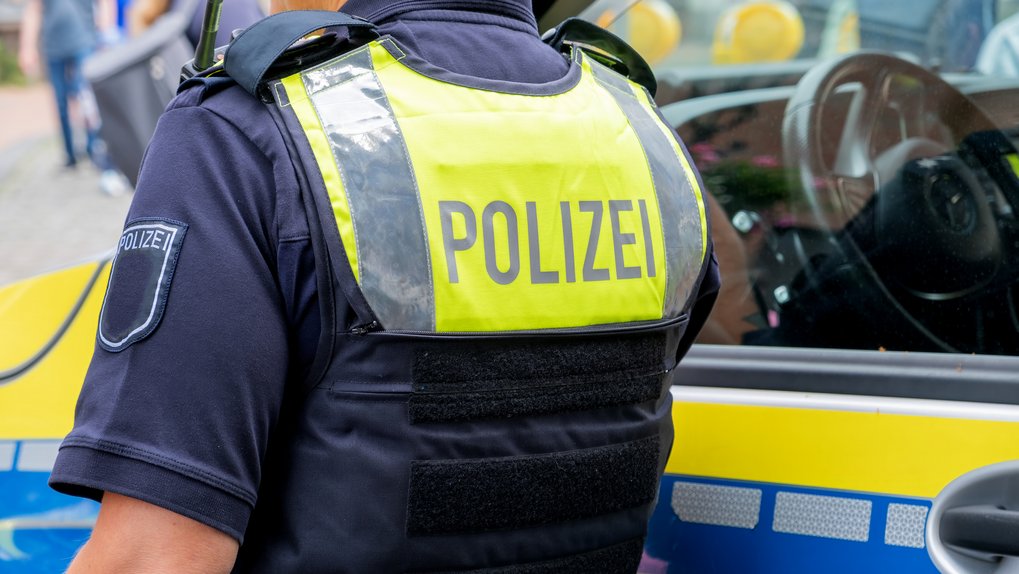 Police officer seen from behind, wearing uniform and yellow vest with police lettering; he is standing next to the police car.