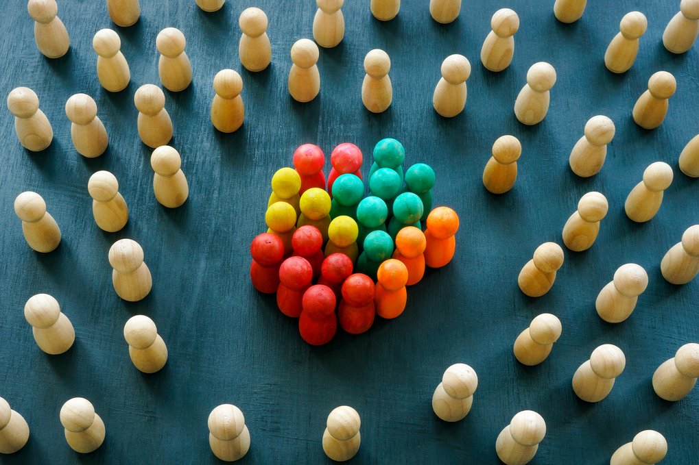 Bunte Figuren sind als Symbol der Inklusivität von einfarbigen Holzfiguren umgeben. (Eine Gruppe bunter Spielfiguren steht in der Mitte, umgeben von beigen Figuren auf einem blauen Untergrund, die einen Kreis bilden.)